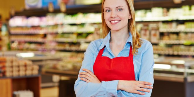 Junge Verkäuferin mit Schürze steht lächelnd im Supermarkt
